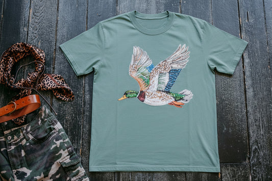 Green t-shirt with a duck graphic on a wooden surface with camouflage pants and a hat.